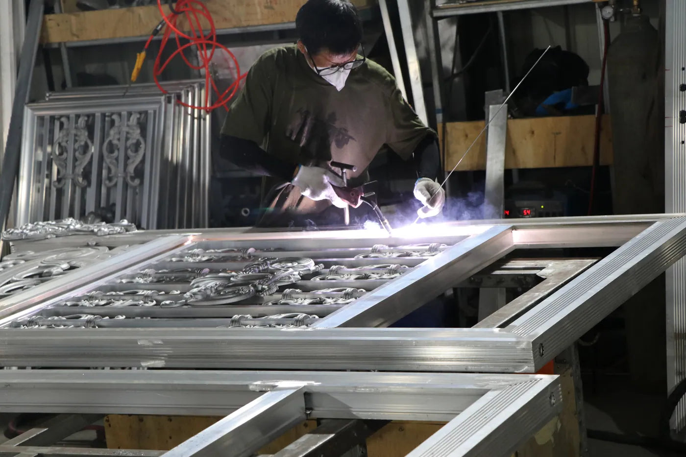 A worker in a mask welding a cast ornamental panel into an aluminium gate frame laid flat on the bench, lit by the arc.