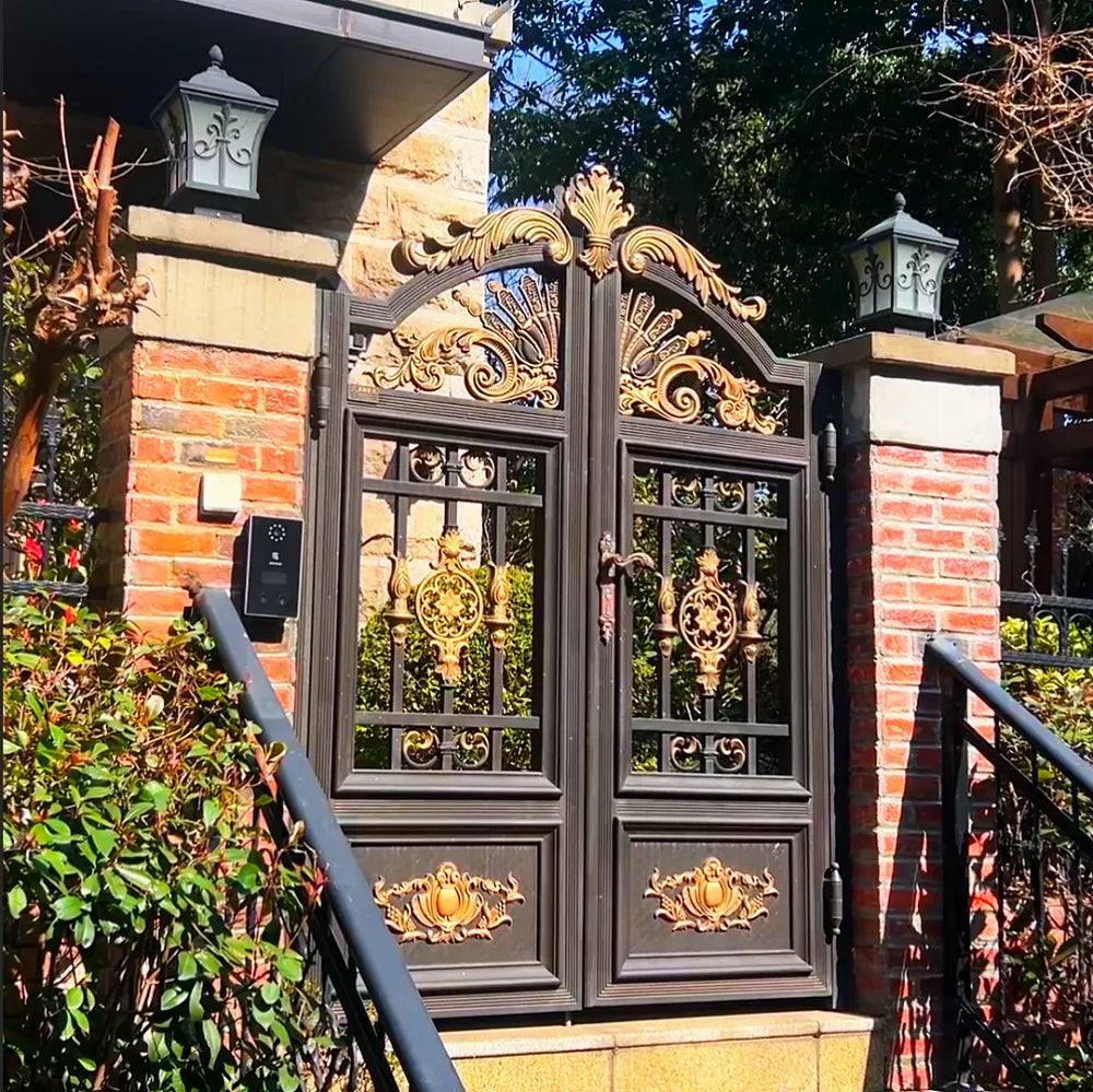 Installation photograph supplied by the works: Bronze aluminium double gate with a gilded cast crown and rosettes, between red brick piers carrying wall lanterns, beside a stepped path with a metal handrail.
