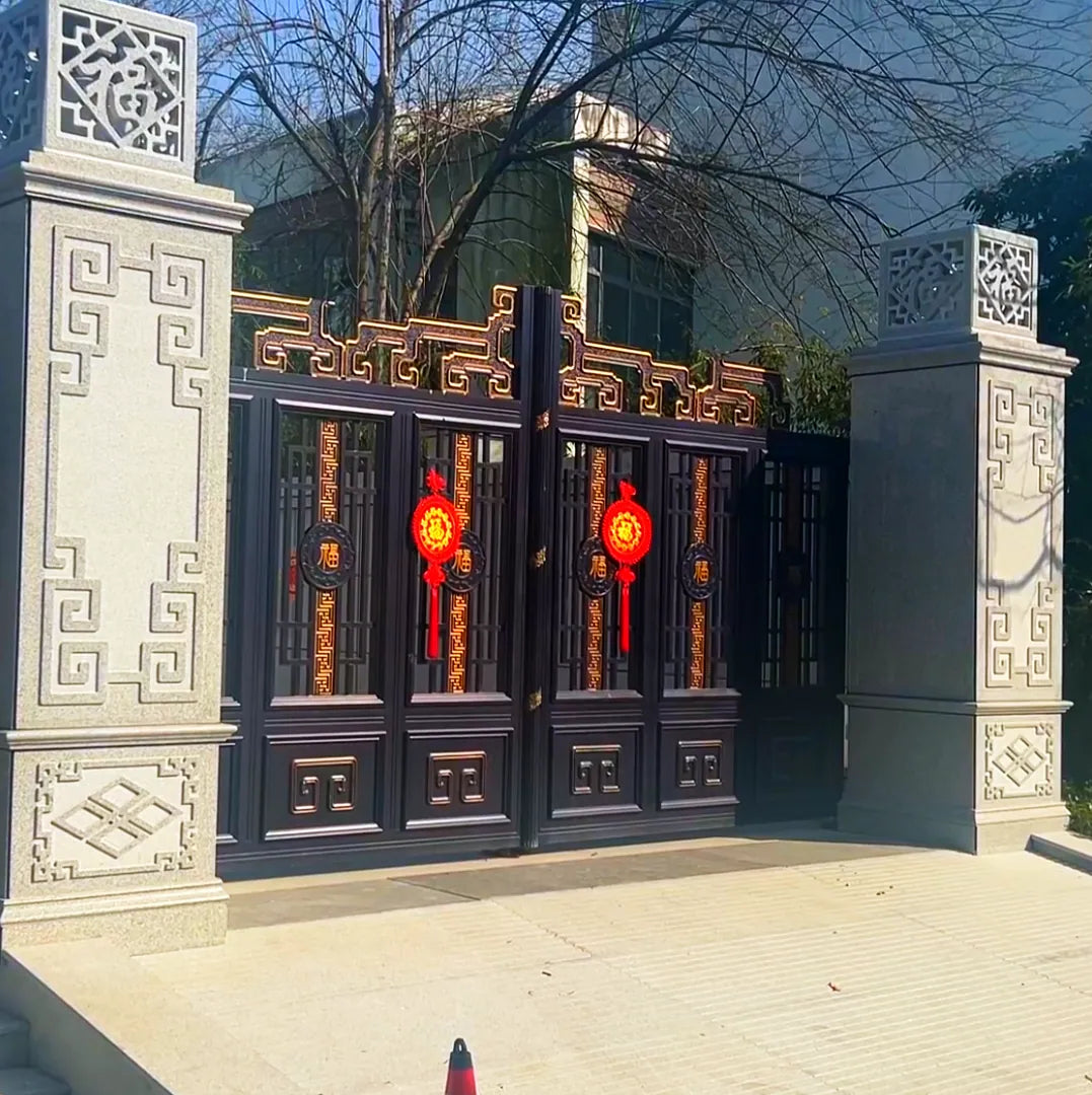 Installation photograph supplied by the works: Black aluminium double gate with fretwork panels and red hanging ornaments, set between carved pale stone piers along a street elevation.