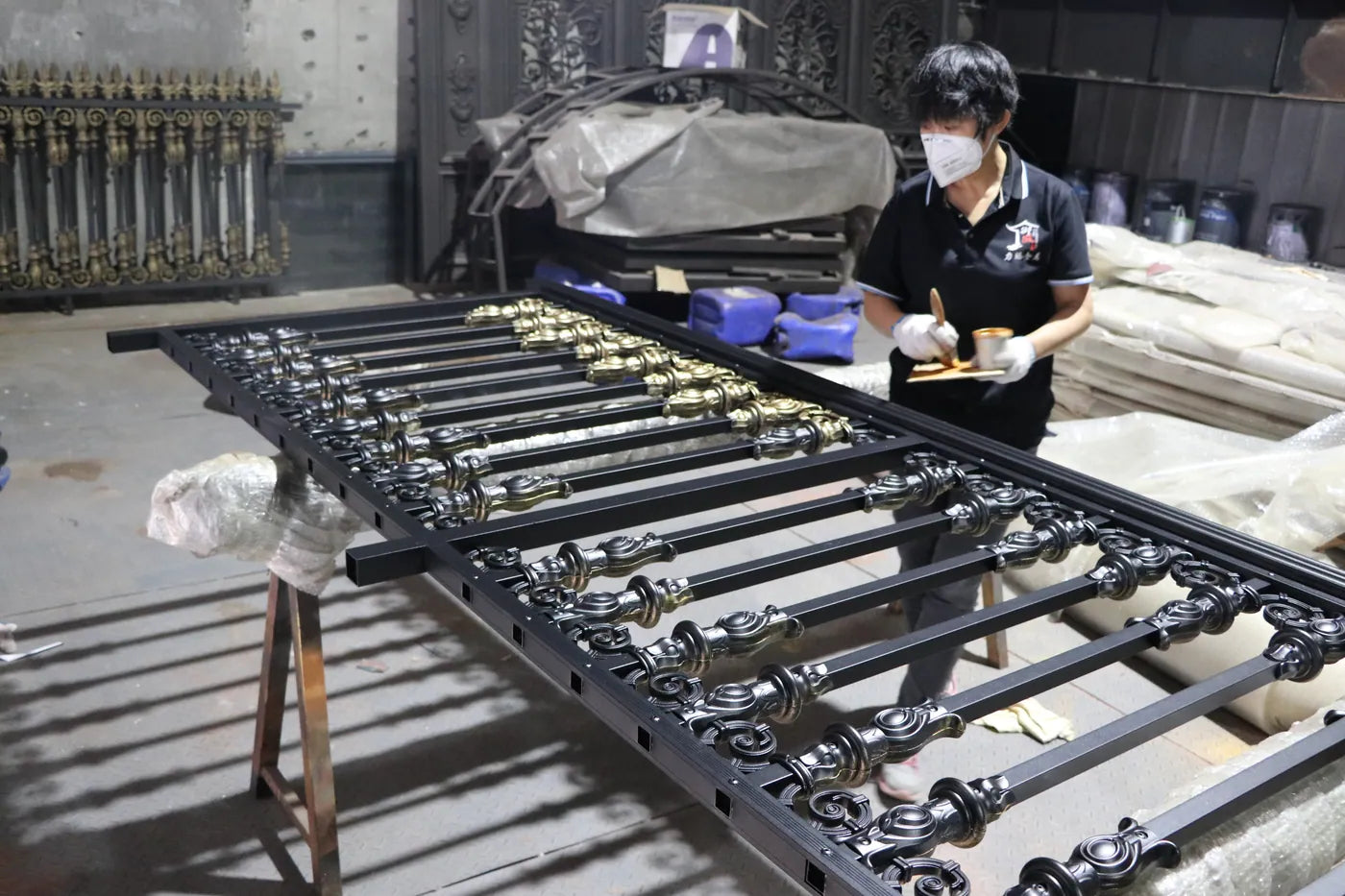 A worker finishing a black gate leaf laid on trestles, its cast ornaments picked out in gold, with more gate parts stacked behind.