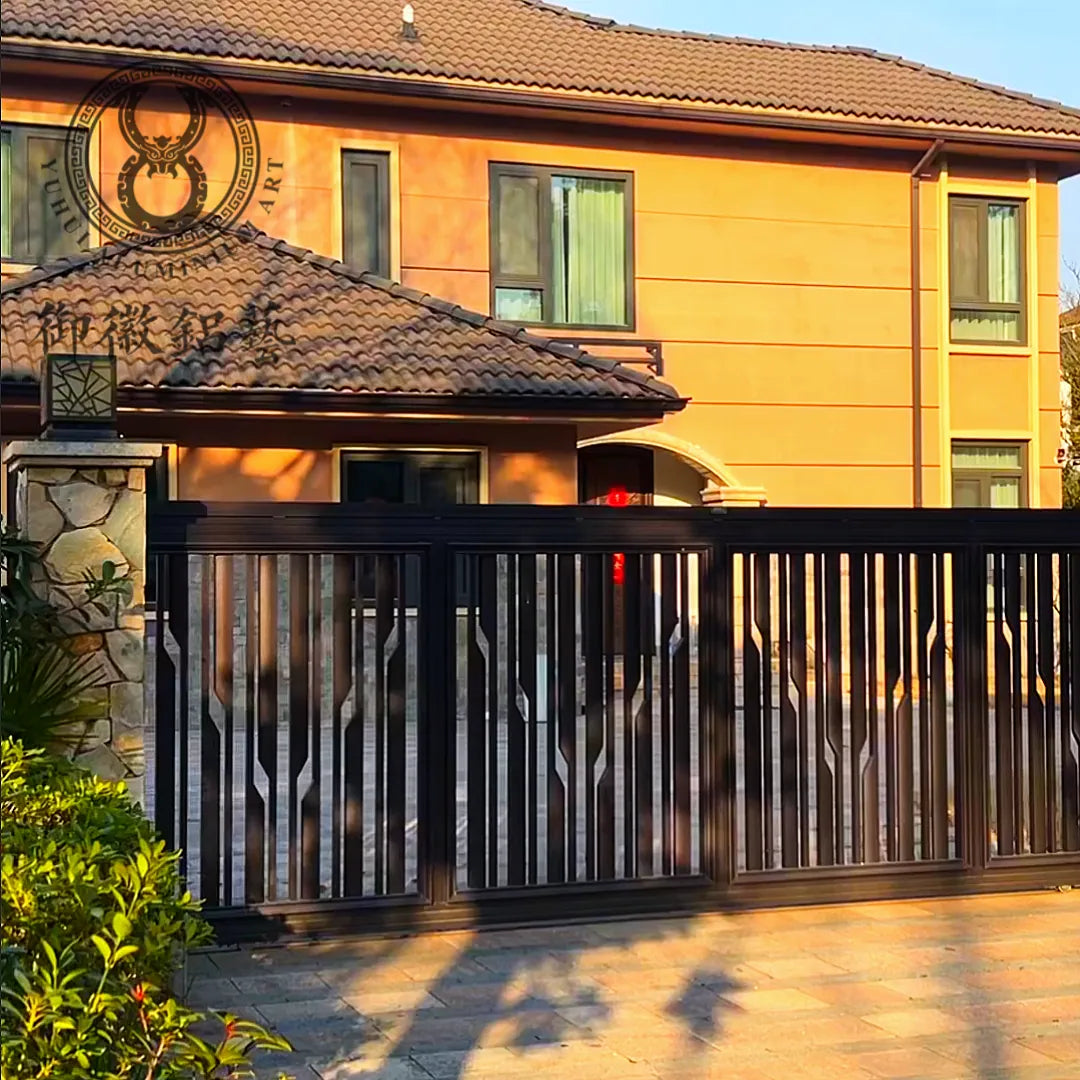 Dark aluminium fence run of tapered vertical blades with a sliding gate leaf, along the front of a tiled-roof house