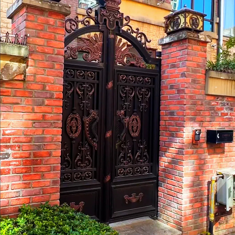 Dark bronze aluminium double gate with dense cast relief panels and an arched cast head, set between red brick piers